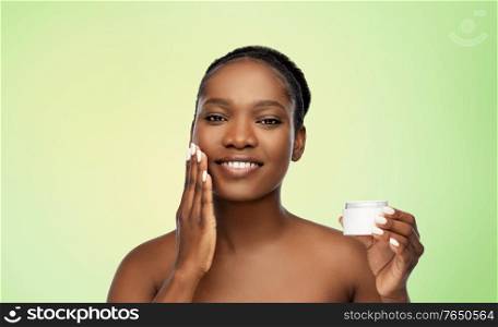 beauty and people concept - portrait of happy smiling young african american woman with bare shoulders applying moisturizer to her face over lime green natural background. smiling african american woman with moisturizer