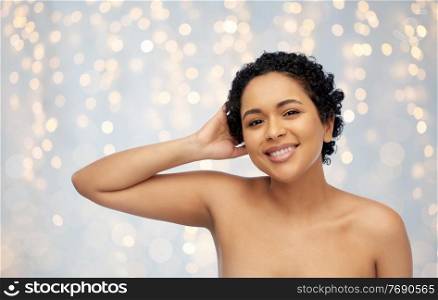 beauty and people concept - portrait of happy smiling young african american woman with bare shoulders touching her hair over festive lights on grey background. portrait of young african american woman
