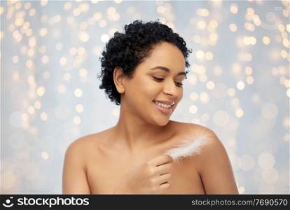 beauty and people concept - portrait of happy smiling young african american woman with bare shoulders and feather over festive lights on grey background. happy african american woman with feather