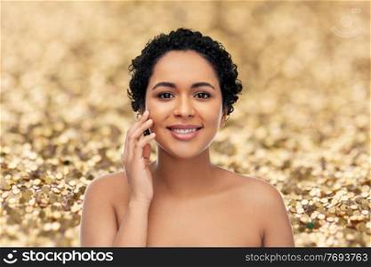beauty and people concept - portrait of happy smiling young african american woman with bare shoulders touching her face over golden glitter background. portrait of young african american woman