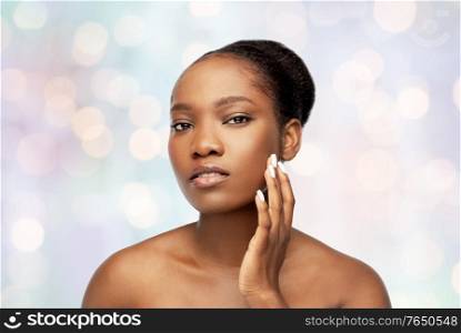 beauty and people concept - portrait of young african american woman with bare shoulders touching her face over holidays lights on blue background. portrait of african woman touching her face