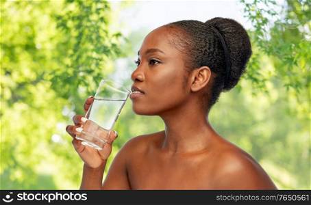 beauty and people concept - portrait of young african american woman with bare shoulders drinking water from glass over green natural background. young african american woman with glass of water