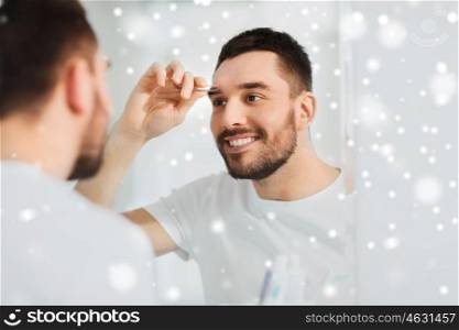 beauty and people concept - smiling young man with tweezers tweezing eyebrow and looking to mirror at home bathroom over snow. man with tweezers tweezing eyebrow at bathroom