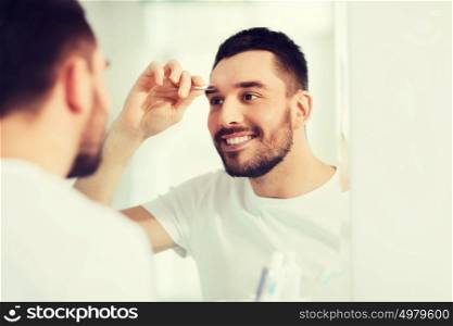 beauty and people concept - smiling young man with tweezers tweezing eyebrow and looking to mirror at home bathroom. man with tweezers tweezing eyebrow at bathroom