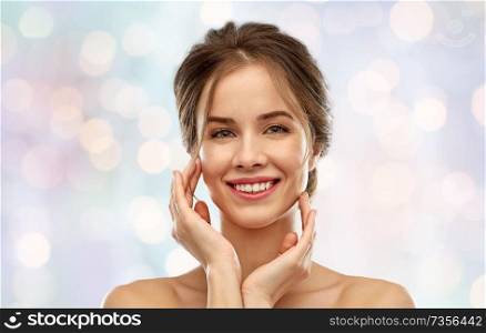 beauty and people concept - smiling young woman touching face over festive lights background. smiling young woman touching her face
