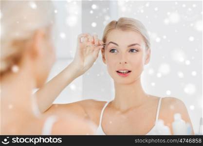 beauty and people concept - smiling young woman with tweezers tweezing eyebrow and looking to mirror at home bathroom over snow. woman with tweezers tweezing eyebrow at bathroom
