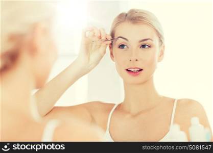 beauty and people concept - smiling young woman with tweezers tweezing eyebrow and looking to mirror at home bathroom. woman with tweezers tweezing eyebrow at bathroom