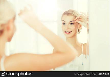 beauty and people concept - smiling young woman with tweezers tweezing eyebrow and looking to mirror at home bathroom. woman with tweezers tweezing eyebrow at bathroom. woman with tweezers tweezing eyebrow at bathroom