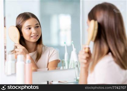 beauty and people concept - teenage girl looking to mirror and brushing hair with comb at home bathroom. teenage girl brushing hair with comb at bathroom