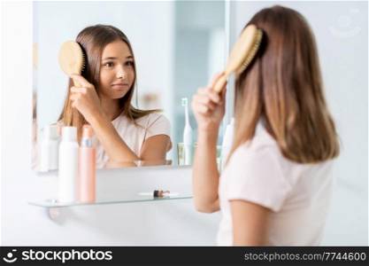 beauty and people concept - teenage girl looking to mirror and brushing hair with comb at home bathroom. teenage girl brushing hair with comb at bathroom