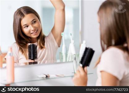 beauty and people concept - teenage girl looking to mirror and singing to hairbrush at bathroom. teenage girl singing to hairbrush at bathroom