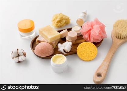 beauty and spa concept - close up of konjac sponge, crafted soap bars, body butter, wisp and natural bristle bath brush and moisturizer on wooden tray. crafted soap, sponges, brush and natural cosmetics