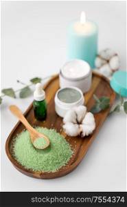 beauty and spa concept - green bath salt, serum with dropper, blue clay mask, moisturizer and eucalyptus cinerea with cotton flowers on wooden tray. bath salt, serum, clay mask and cotton on tray