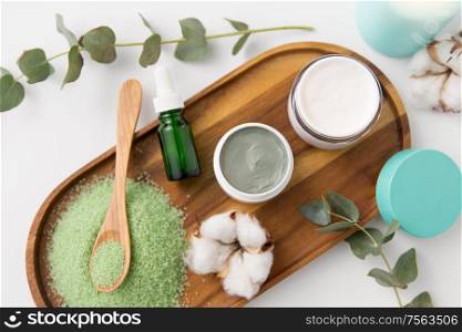 beauty and spa concept - green bath salt, serum with dropper, blue clay mask, moisturizer and eucalyptus cinerea with cotton flowers on wooden tray. bath salt, serum, moisturizer and oil on tray