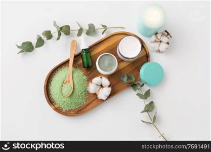 beauty and spa concept - green bath salt, serum with dropper, blue clay mask, moisturizer and eucalyptus cinerea with cotton flowers on wooden tray. bath salt, serum, moisturizer and oil on tray