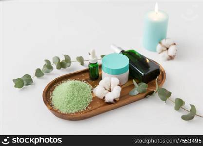 beauty and spa concept - green bath salt, serum with dropper, body oil, moisturizer and eucalyptus cinerea with cotton flowers on wooden tray. bath salt, serum, moisturizer and oil on tray