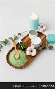 beauty and spa concept - green bath salt, serum with dropper, moisturizer, blue clay mask and eucalyptus cinerea with cotton flowers on wooden tray. bath salt, serum, moisturizer and oil on tray