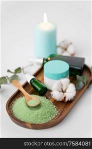 beauty and spa concept - green bath salt, serum with dropper or essential oil, moisturizer and eucalyptus cinerea with cotton flowers on wooden tray. bath salt, serum, moisturizer and oil on tray