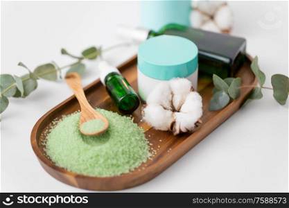 beauty and spa concept - green bath salt, serum with dropper or essential oil, moisturizer and eucalyptus cinerea with cotton flowers on wooden tray. bath salt, serum, moisturizer and oil on tray