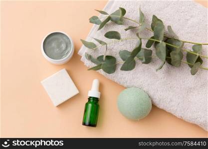 beauty and spa concept - serum or essential oil, clay mask, soap bar, konjac sponge and eucalyptus cinerea on bath towel. serum, clay mask, oil and eucalyptus on bath towel