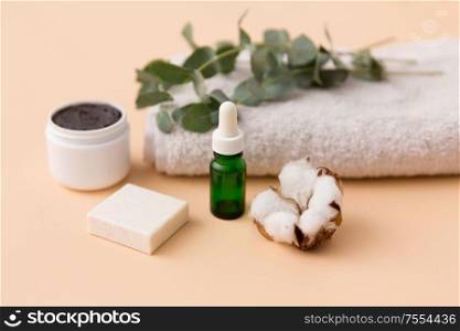 beauty and spa concept - serum or essential oil, mask, soap bar, eucalyptus cinerea and cotton flower on bath towel. serum, clay mask, oil and eucalyptus on bath towel