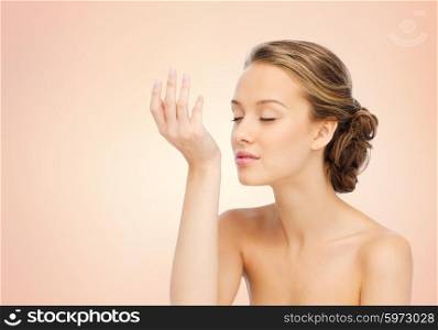 beauty, aroma, people and body care concept - young woman smelling perfume from wrist of her hand over beige background