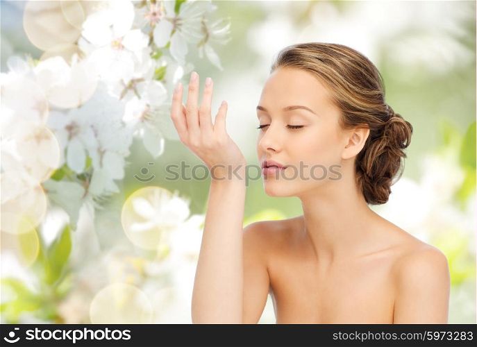 beauty, aroma, people and body care concept - young woman smelling perfume from wrist of her hand over green natural background with cherry blossoms