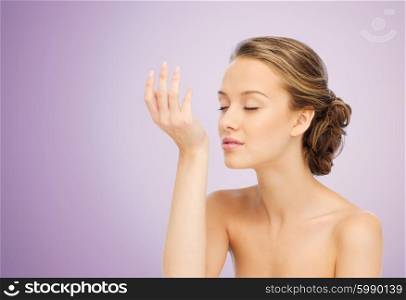 beauty, aroma, people and body care concept - young woman smelling perfume from wrist of her hand over violet background