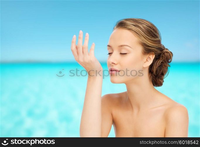 beauty, aroma, people and body care concept - young woman smelling perfume from wrist of her hand over blue sea and sky background