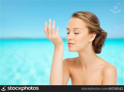 beauty, aroma, people and body care concept - young woman smelling perfume from wrist of her hand over blue sea and sky background