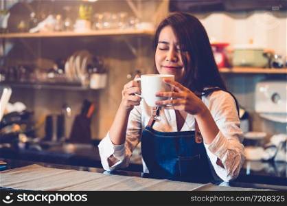 Beauty Asian female barista holding cup of coffee and tasting brewed coffee with cafe restaurant background. Waitress smelling coffee at cafeteria. Food and drink concept. People lifestyle