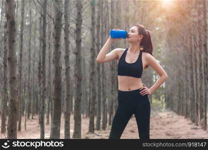 Beauty Asian sport woman resting and holding drinking water bottle and relaxing in middle of forest after tired from jogging. Girl looking attraction view. Workout concept. Lifestyle theme