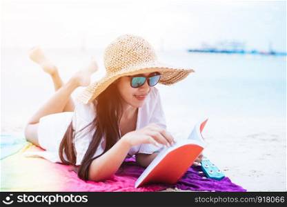 Beauty Asian woman have vacation on beach. Girl wearing wing hat and reading book on colorful mat near sea. Lifestyle and happy life concept. Travel and holiday theme.