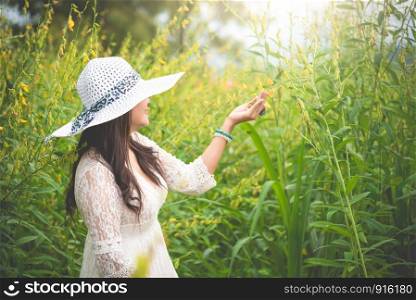 Beauty Asian woman in white dress and wing hat walking in rapeseed flower field background. Relaxation and travel concept. Wellness happy life of girl on vacation. Nature and people lifestyle