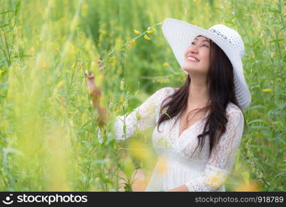 Beauty Asian woman in white dress and wing hat walking in rapeseed flower field background. Relaxation and travel concept. Wellness happy life of girl on vacation. Nature and people lifestyle
