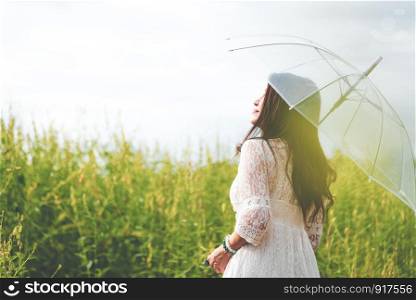 Beauty Asian woman in white dress holding transparent umbrella and look at sky between rapeseed flower field background. People relaxation and travel concept. Wellness happy life of girl on vacation.