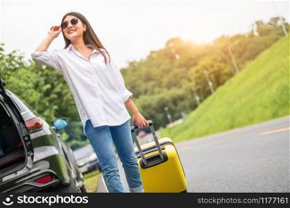 Beauty Asian woman with sunglasses dragging yellow suitcase baggage alone on road trip. People lifestyle and vacation concept. Nature and summer background. Girl having car adventure transportation