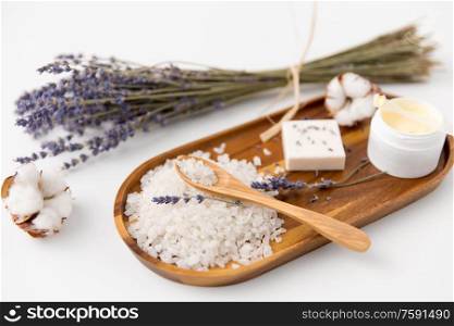 beauty, bath and wellness concept - sea salt with spoon, soap bar, body butter, lavender and cotton flowers on wooden tray. bath salt, lavender soap and body butter on tray