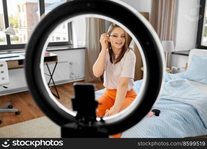 beauty blogging, make up and people concept - happy smiling girl blogger with ring light and smartphone applying mascara at home. girl blogger with ring light applying make up