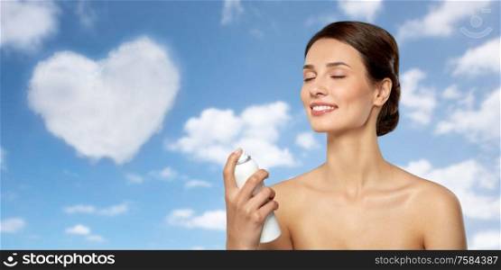 beauty, bodycare and people concept - beautiful young woman applying facial spray or mist to her face skin over blue sky and heart shaped cloud background. beautiful young woman with facial spray or mist