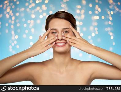 beauty, bodycare and people concept - beautiful young woman touching her face over holidays lights on blue background. beautiful young woman touching her face
