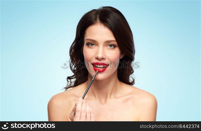 beauty, cosmetics and people concept - beautiful young woman with make up brush applying red lipstick over blue background. beautiful woman with make up brush for lipstick