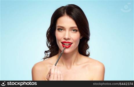 beauty, cosmetics and people concept - beautiful young woman with make up brush applying red lipstick over blue background. beautiful woman with make up brush for lipstick