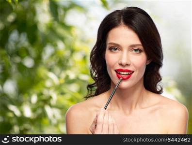 beauty, cosmetics and people concept - beautiful young woman with make up brush applying red lipstick over green natural background. beautiful woman with make up brush for lipstick