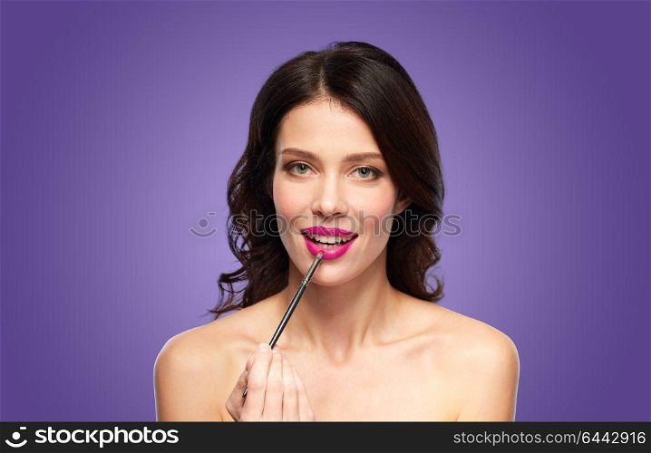 beauty, cosmetics and people concept - beautiful young woman with make up brush applying berry lipstick over ultra violet background. beautiful woman with make up brush for lipstick