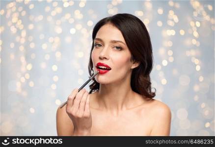 beauty, cosmetics and people concept - beautiful young woman with make up brush applying red lipstick over holidays lights background. beautiful woman with make up brush for lipstick. beautiful woman with make up brush for lipstick