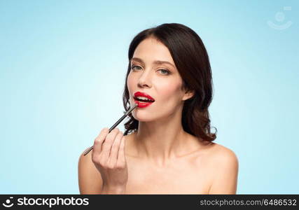 beauty, cosmetics and people concept - beautiful young woman with make up brush applying red lipstick over blue background. beautiful woman with make up brush for lipstick. beautiful woman with make up brush for lipstick