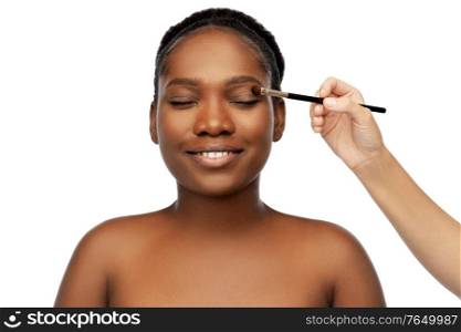 beauty, cosmetics and people concept - portrait pf of beautiful smiling young african american woman and hand with make up brush applying eye shadow over white background. face of african woman and hand with make up brush