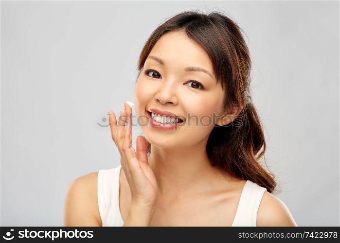 beauty, cosmetics and skincare concept - happy young asian woman applying moisturizer cream to her face over grey background. young asian woman applying moisturizer to her face