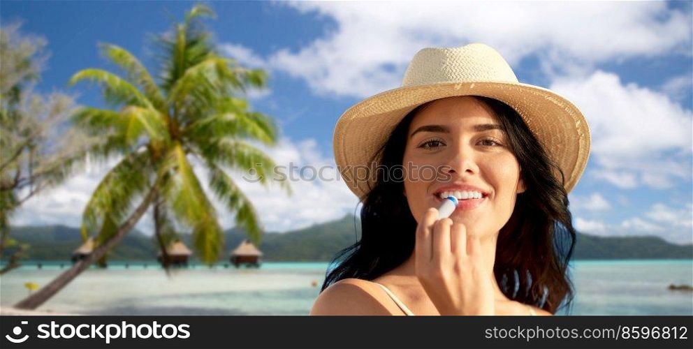 beauty, cosmetics and summer holidays concept - portrait of happy smiling young woman in bikini swimsuit and straw hat applying lip balm over tropical beach background in french polynesia. smiling woman in bikini with lip balm on beach